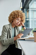 © WHstudio Leushin N - Indoor shot of serious curly haired successful businesswoman concentrated in tablet watches video online pose near windowsill in coworking space writes down notes to remember important information