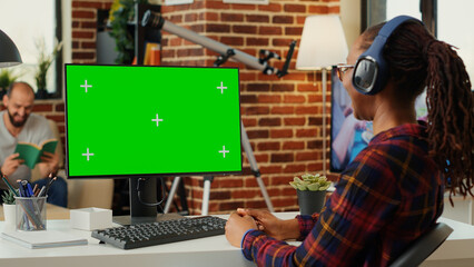 African american woman working with greenscreen and listening to song on headphones, using computer with chroma key display and isolated mockup. Looking at blank green screen.