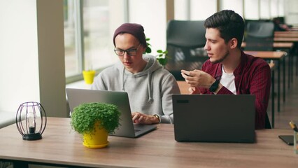 Two hipster coworkers sharing ideas about project sitting with laptops in spacious open space office. Developer discuss software program code. Designer and manager consulting on working plan.