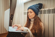 © Westend61 - Woman wearing knit hat using laptop leaning on radiator at home