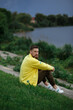 © Iryna Savchuk - 28.09.22 Vinnytsia -Ukraine: A bearded guy is sitting on the grass in a yellow jacket near the lake at sunset. There should be a thunderstorm and a blue rainy sky