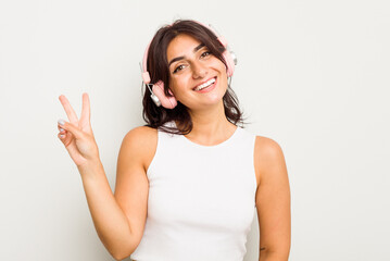 Wall Mural - Young Indian woman wearing headphones isolated on white background joyful and carefree showing a peace symbol with fingers.