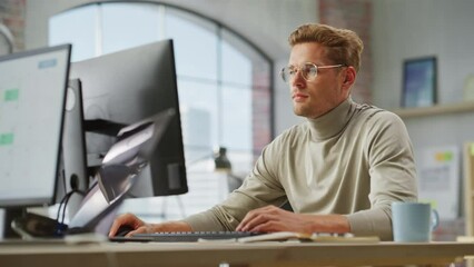 Poster - Portrait of White Creative Young Man With Glasses Working on a Computer During Day Time in a Spacious Modern Office. Male Graphic Designer Smiling While Developing a New Visual Concept. Dolly Shot.