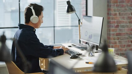 Fototapete - Portrait of Asian Creative Young man Smiling While Using Computer in Modern Office. Logistics Supervisor Putting Headphones on, Starts Analyzing Charts, Stock Market, Financial Data Shown on Display