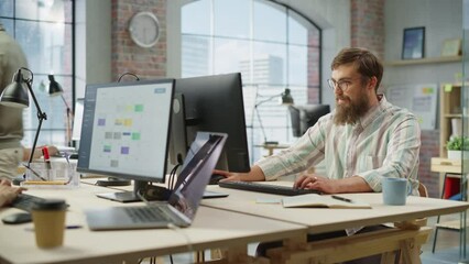 Poster - Portrait of Creative Bearded White Man with Glasses Smiling While Using Computer in Bright Spacious Office. Male Team Manager Giving Feedback to colleagues. People Working in Background. Dolly Shot.