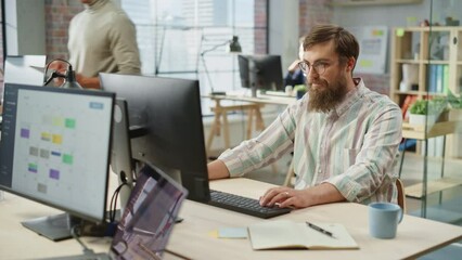 Poster - Portrait of Young Caucasian Bearded Man with Glasses Smiling While Sitting and Working on Computer in a Bright Spacious Office. Male Legal Consultant Typing Notes on Keyboard. Dolly Shot.