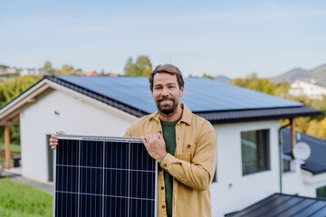 Mature man holding solar panel near his house with solar panels on the roof. Alternative energy, saving resources and sustainable lifestyle concept.