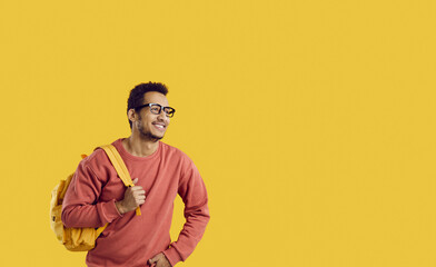 Happy cheerful college or university student with backpack isolated on copy space background. Handsome young black man in sweatshirt and glasses smiling and looking away at yellow studio background