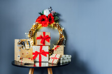 Various Christmas Gift Boxes On The Black Table With Elegant Christmas Flower Wreath On Gray Wall Background. Luxury Gifts Packing With Golden Boxes And Red Ribbons. Festive Concept. Selective Focus