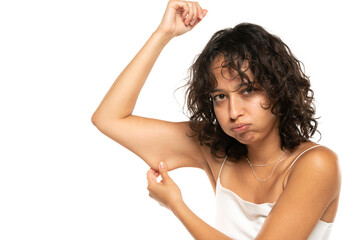 Wall Mural - Unhappy etnic woman testing the flabby muscle under her arm pulling it down with her hand as she checks for muscle tone or weight gain on a white background