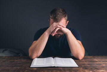 Wall Mural - A man prays with folded hands. An open Holy Bible on the table. A young man with a beard.