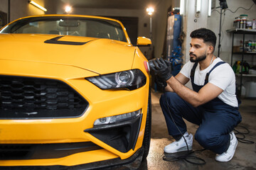 Car detailing and polishing concept. Professional young male car service worker in uniform, holding in hands orbital polisher, and polishing yellow luxury car at auto repair service station.