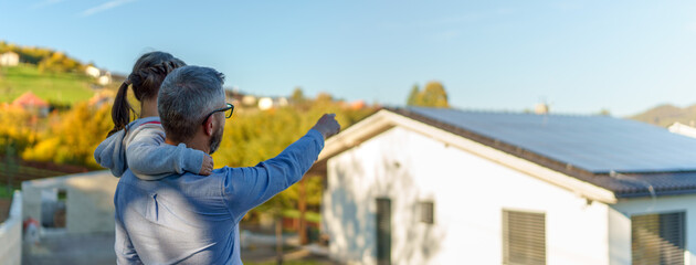 Wall Mural - Rear view of dad holding her little girl in arms and showing at their house with installed solar panels. Alternative energy, saving resources and sustainable lifestyle concept.