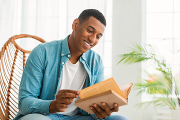 Smiling smart millennial african american guy reading book, sit on chair, enjoy comfort and free time