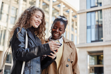 Waist up portrait of two smiling young women using smartphone while standing outside shopping mall, African American and Caucasian