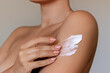Cropped shot of a young tanned woman moisturizing her shoulder with her hand using a cream isolated on a gray background. Skin care. A white thick smear of cream on the female body