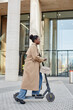 Vertical side view portrait of young black woman riding electric scooter in city and smiling cheerfully while wearing long trenchcoat