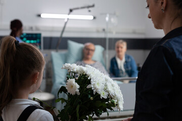 Wall Mural - Unrecognizable girl holding grandfather flowers while frican american doctor performs routine medical checkup. Grandparents greeting daughter, granddaughter in elderly man clinic recovery room.