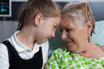 Wall Mural - Elderly woman lying in hospital bed with breathing aids showing affection to little girl. Grandmother and granddaughter bumping heads in love at medical clinic for elderly people recovery room.