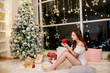 Xmas tree decorated by lights gifts toys, candles, lanterns, garland lighting indoors holiday living room new year. Asian woman in white sweater, warm and cozy evening in Christmas interior design.
