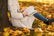 © Prostock-studio - Young european red-haired female in raincoat reads book, sit near tree in park with yellow leaves