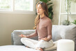 © KMPZZZ - Air humidifier, calm asian young woman sitting on couch lotus pose put hands practice meditation do yoga exercise at home. Aromatherapy steam scent from essential oil diffuser in living room at home.