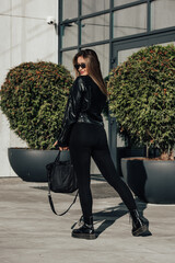 Wall Mural - Fashionable young woman with long hair wearing solid long sleeve bodycon one piece jumpsuits posing in a black leather jacket and smooth platform boots