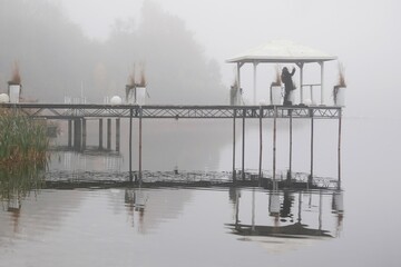 Poster - Romantic moody scenery with wooden pier with white gazebo and plants in white flower pots by lake on calm misty day. Silhouette of woman taking photo on pier.