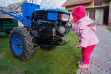 Little Girl Near A Tractor,a Baby Looks At A Two-wheeled Tractor In The Yard
