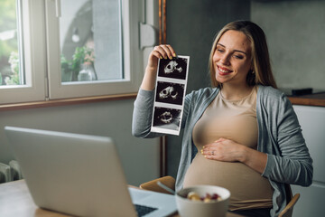 side view of caucasian pregnant woman showing ultrasound scanner picture while having video call in her kitchen