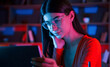 © standret - Focused on the work. Beautiful woman in glasses and red wear is sitting by the laptop in dark room with neon lighting