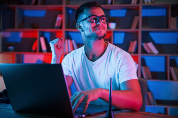 Positive emotions. Man in glasses and white shirt is sitting by the laptop in dark room with neon lighting