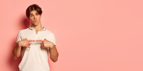 Wall Mural - Portrait of young man in casual white T-shirt posing with shy expression over pink background. Flyer. Love