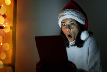 Woman In Santa Hat Holding Digital Tablet In Her Hands And Looking At Screen In Surprise With Wide Eyes.