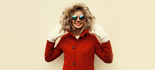 Wall Mural - Portrait of happy smiling young woman wearing red jacket with fur hood and mittens on background