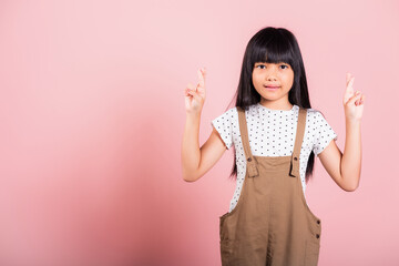 Asian little kid 10 years old smiling crossing fingers at studio shot isolated on pink background, Portrait of Happy child girl wishing for good luck, hoping her wish come true luck and superstition