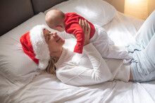 Happy Mother Lies On Bed In Red Santa Hat And White T-shirt, Holds On Hands Baby In Red T-shirt And Smile. Newborn - Happiness For Family. New Year And Christmas. Elf And Fairy. Side View.