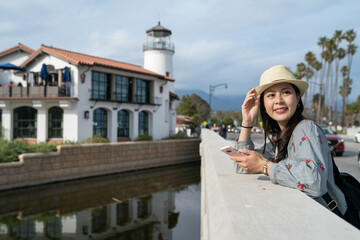 Canvas Print - leisure asian Chinese female traveler holding hat leaning on stone railing and looking into distance at nice view in santa Barbara with a lighthouse at background at dusk