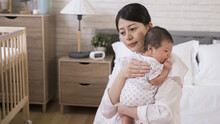 Portrait Asian Mother Is Helping Her Adorable Newborn To Burp With Gentle Pat On The Back In The Bedroom At Home.