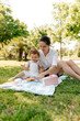 European woman with long dark hair wearing white shirt and shorts sitting on blanket in the park with her little daughter and having picnic in sunny day. 