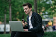© Iona - Handsome brunette cheerful male sitting on grass at park with laptop making video call holds phone smiling having break remote working. Cheerful man student talking with friend via internet.