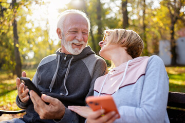 Wall Mural - Cheerful seniors in sports clothing sitting on a park bench after exercise looking at smartphones, talking, and smiling. An active elderly couple in the park using a smartphone