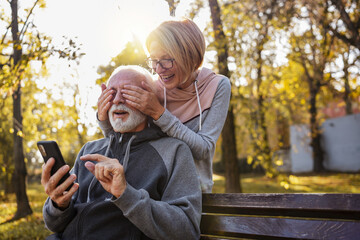 Wall Mural - An active modern cheerful senior couple meets in the park. An older man is sitting on a park bench using a smartphone, and an elderly woman surprises him from the back.