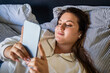 European woman wearing earphones laying at the bed and looking at her cellphone