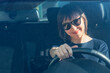 Woman sits in a car in sunglasses, holds the steering wheel and smiles