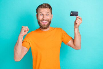 Wall Mural - Photo of positive impressed man with masculine beard dressed orange t-shirt hold plastic credit card isolated on teal color background