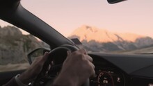 Male Hands Turn Steering Wheel Of Modern Automobile. Man Drives Car On Mountainous Road By Rocky Mountains With Sunlit Peaks. Landscape Of Alps At Dawn Closeup