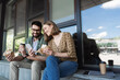 © LIGHTFIELD STUDIOS - Smiling businesswoman showing smartphone to colleague with sandwich and coffee to go outdoors.