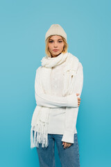 Poster - young blonde woman in white sweater and winter hat standing isolated on blue.