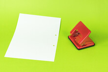 A red metal hole punch and a white sheet of paper on a green background.